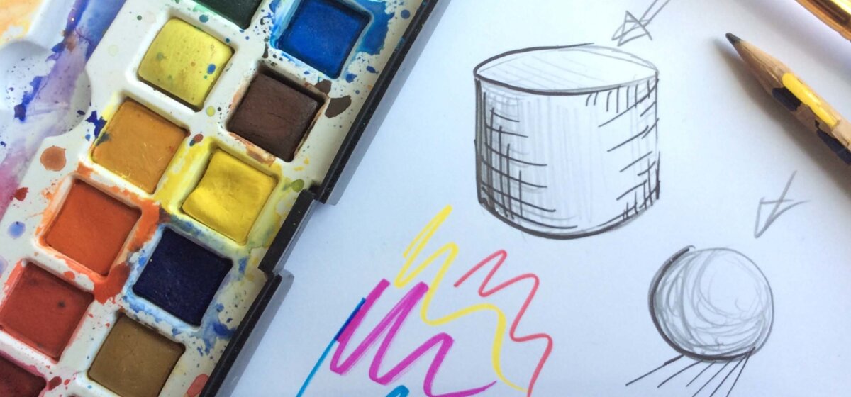 A tray of multicoloured paints sits next to a white canvas, where some shapes have been drawn on in pencil.