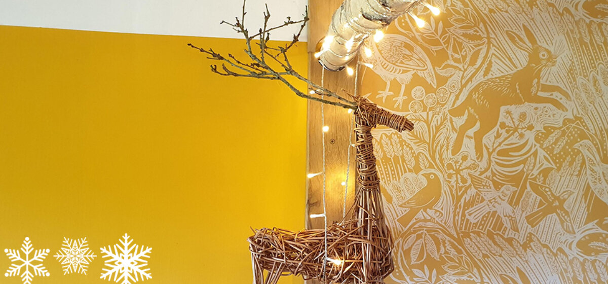 An image of a reindeer made from willow sticks against a yellow background with fairy lights and snowflakes in the corners.