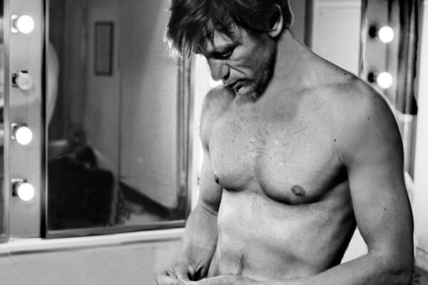 Daniel Craig, the actor, is standing in a room topless looking at his hands.