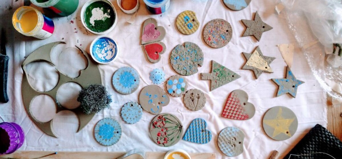 An image for Festive Clay Decoration Making at The Base, Greenham, pictured is various shapes cut out of clay surrounded by paints to decorate them
