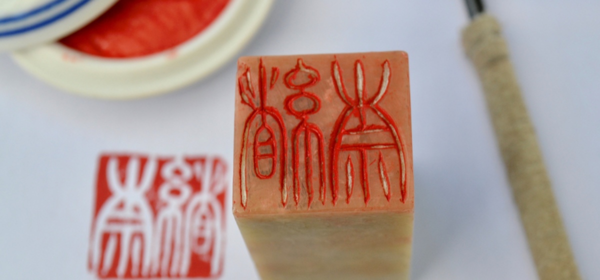 A birds eye image of a square stone stamp with carvings in it and a piece of paper next to it with the print from the carvings on. There is an ink pad in the background.