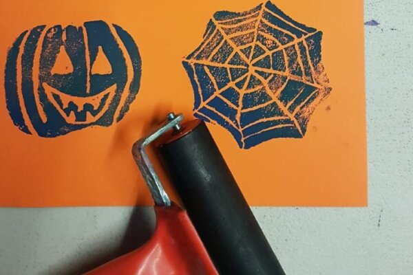 An image for Children's Halloween Printmaking at The Base Greenham, pictured is a orange sheet of paper with a black pumpkin print and a black spiderweb print on it with a roller next to the paper.