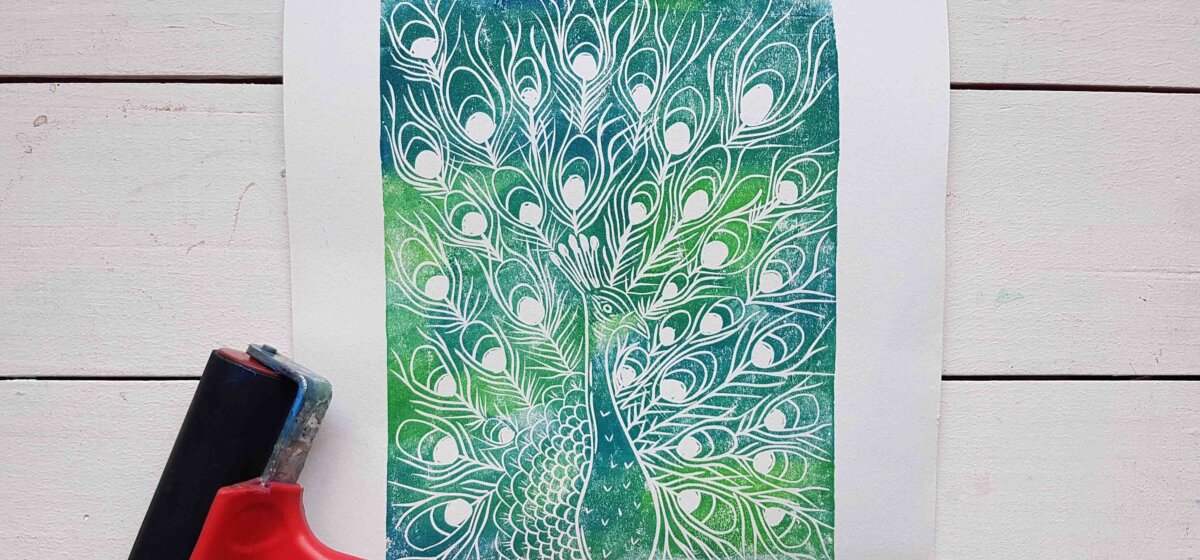 A design of a peacock lineart with a mixture of green and blue colours painted on top. A linoprinting tool features in the bottom left corner.