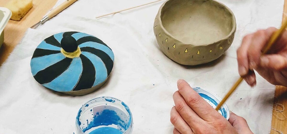 A person holding a paintbrush is decorating a clay pot and lid in blue and black paint.
