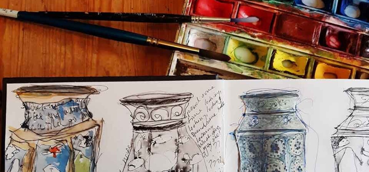 A sketchbook filled with drawings, paintings and written notes is lying on top of a wooden table, next to paintbrushes and a watercolour paint tin.