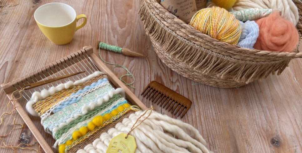 A woven piece made of creams, oranges and blues sits on a wooden table next to a bowl of yarn