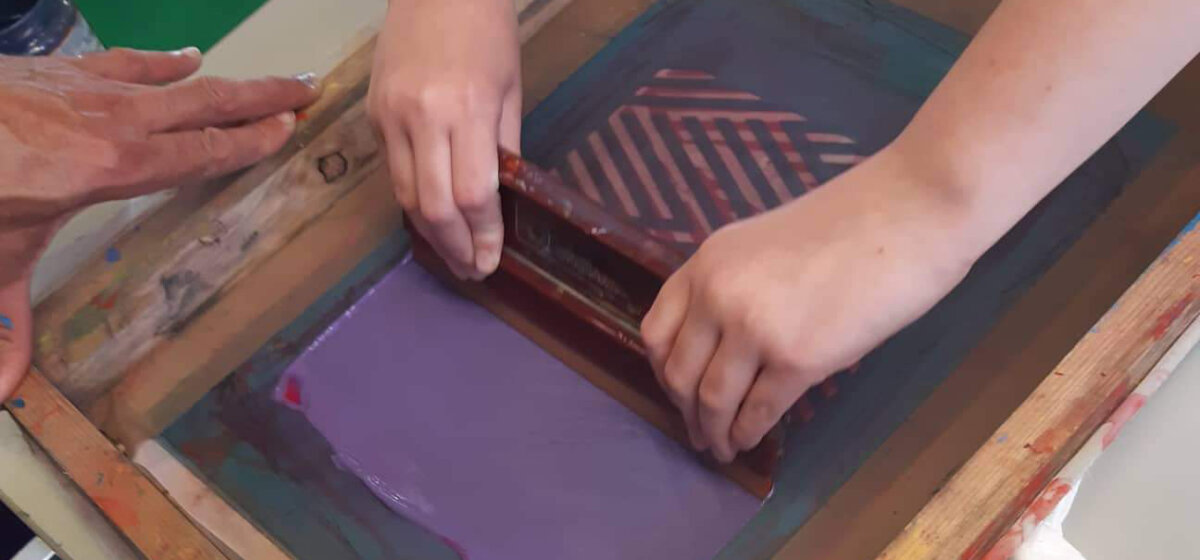 There is a screen printing frame and a young person's hands are squeezing purple paint over their design