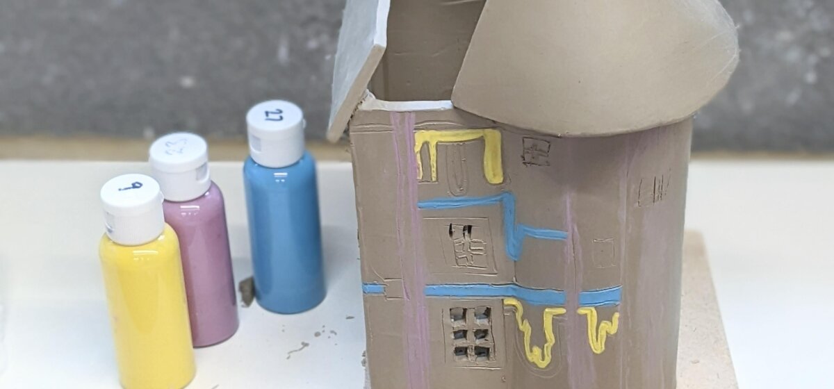 A clay castle with a small amount of paint on it. Next to it are a small line of bottles of paint.