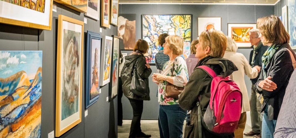 A group of people are standing in an art gallery looking at various art pieces displayed on a wall.