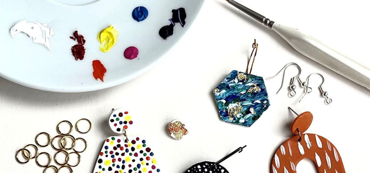 Polymer clay earrings, an assortment of paint and a paint brush are on a white surface.