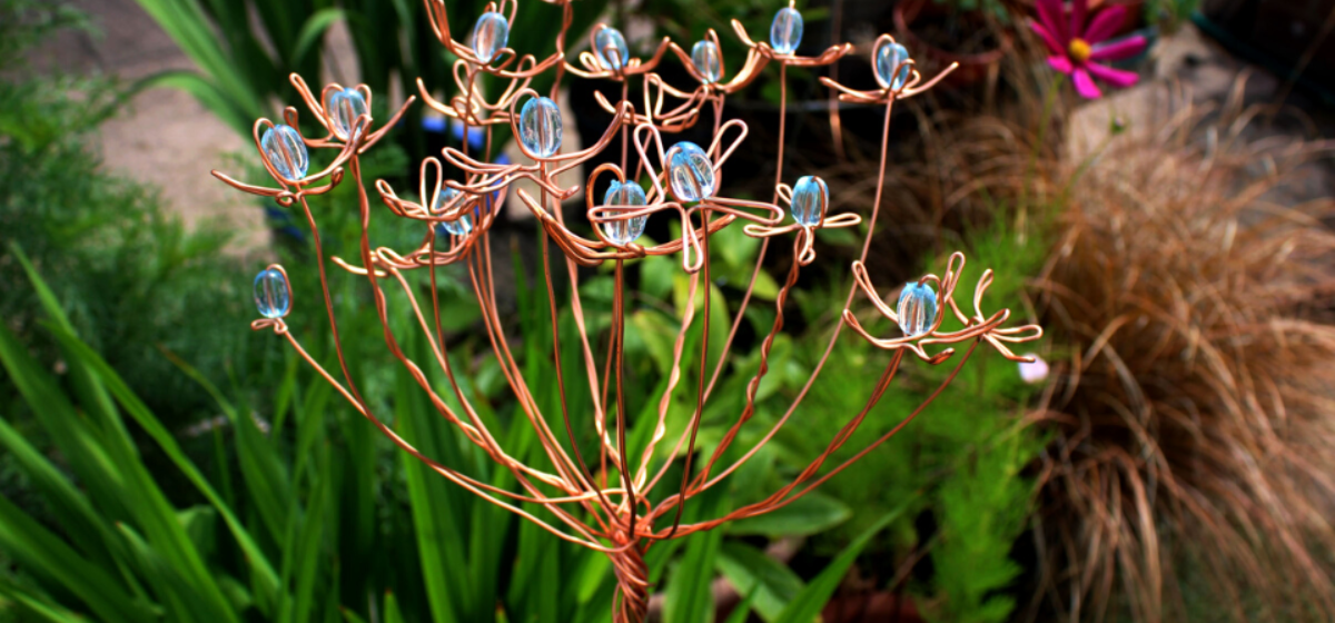 A photo of a garden with a copper wire designed into a flower shape with iridescent beads emulating the middle of a flower.