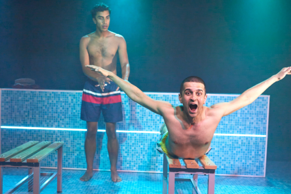 A photo of a stage on which there is a boy laying on his stomach with his arms out in the butterfly swim position on a bench and another boy behind him looking concerned. Behind them both is a swimming pool.