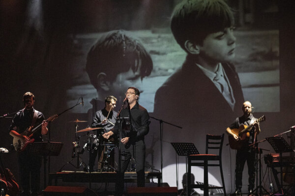 Band on a dark stage, holding instruments with a projection of a black and white photo of children in the background