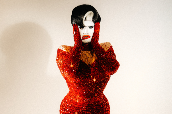A drag queen with white face paint, short black hair and a figure hugging red sequined dress