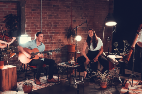 Four people sit in an urban style room, full of exposed brickwork, plants and lamps as they play their instruments