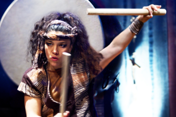 A woman in a striped top and matching head piece and wrist cuffs holds her drum sticks high in the air as she prepares to play.