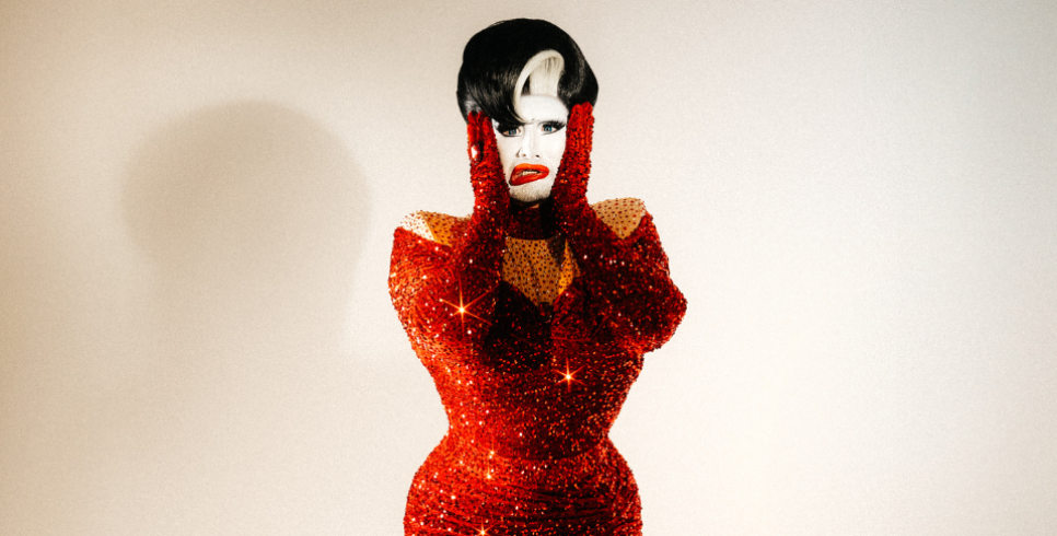 A drag queen with white face paint, short black hair and a figure hugging red sequined dress