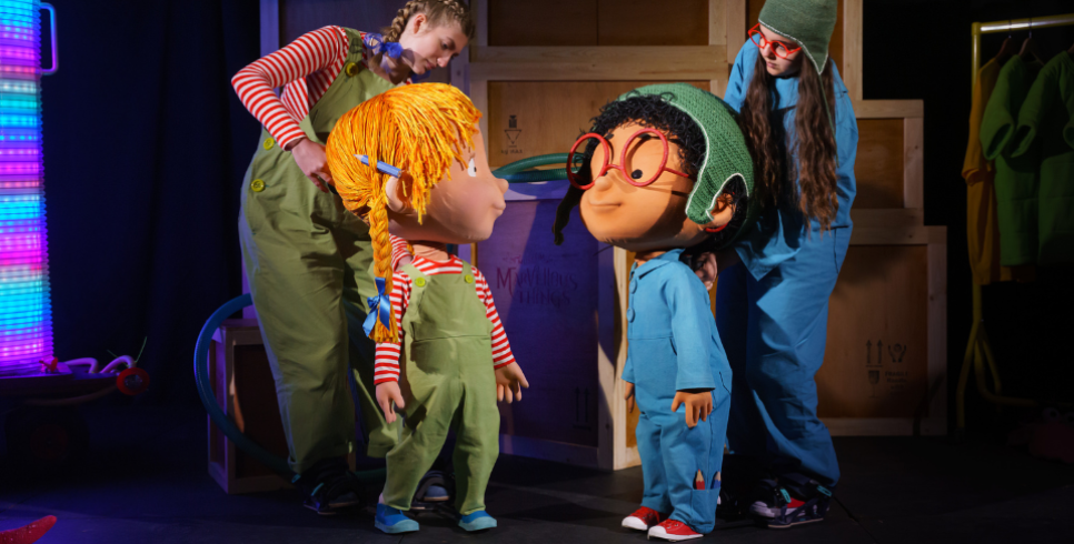 Two puppets on stage, one with orange hair and green dungarees and one with black hair, red glasses and a blue boiler suit.