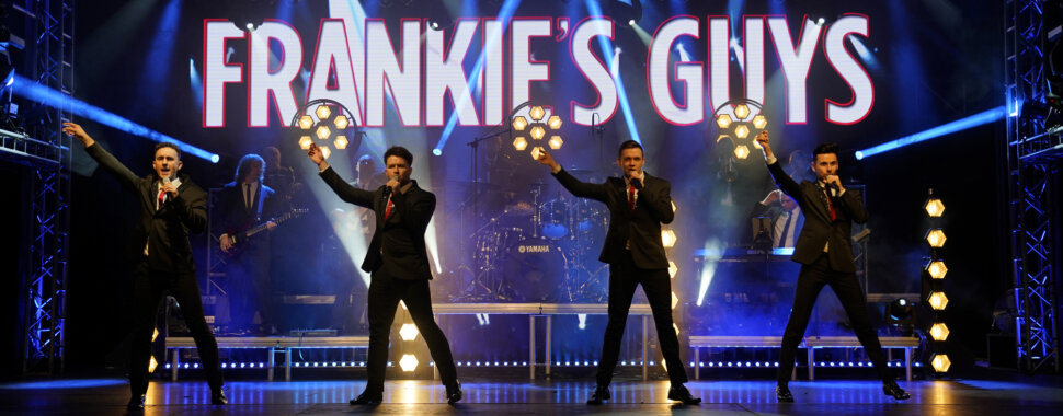 Four men in suits stand with microphones in their hands on a stage lit with a 'Frankie's Guys' sign.