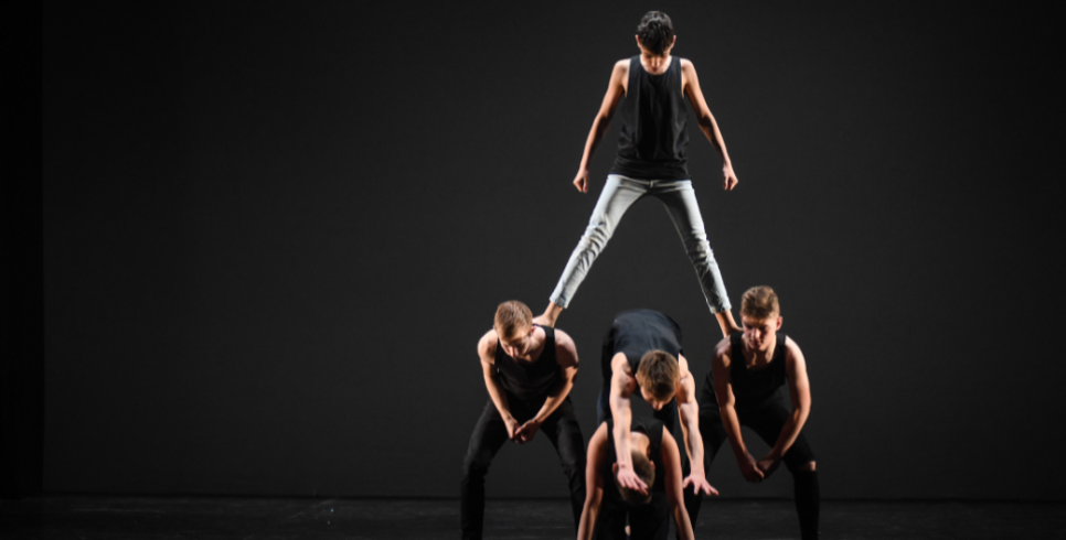 A photograph of a human pyramid made up of 5 young boys. The photo is against a black backdrop.