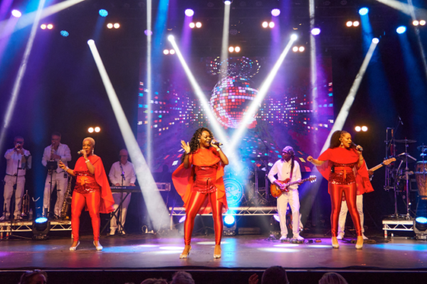 3 black women stand front and centre on stage, singing while wearing full red outfits as they are backed by other musicians as well as colourful lighting