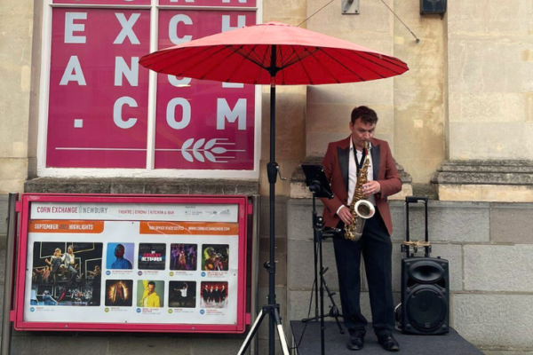 A man in a suit, shaded by an umbrella, stands outside the Corn Exchange playing a saxophone