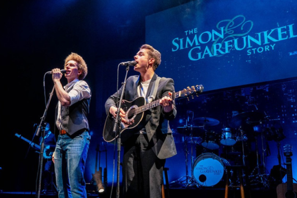 Two men singing into microphones on stage, one playing guitar with a blue background that reads The Simon and Garfunkel Story.