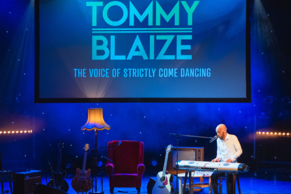 Tommy Blaize performs on stage in a living room-like set, full of cosy lamps and arm chairs on a stage lit in blue light