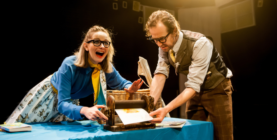 A woman with glasses and in a blue shirt and skirt is pulling an excited face a while she uses an old printing machine. A man with glasses to her right in a shirt and waistcoat is looking down at the machine and assisting.