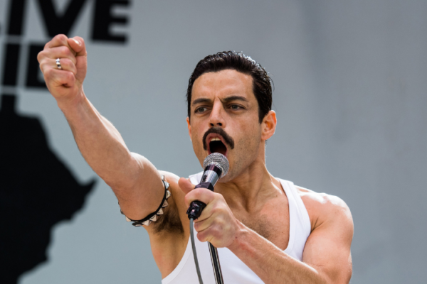 A still from Bohemian Rhapsody of Rami Malek as Freddy Mercury wearing a white tank and singing on a stage with Live Aid behind him.