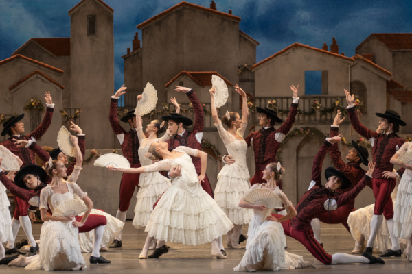 An image of lots of ballet dancers in white layered dresses and white fans and men in burgundy suits with hats dancing in front of a set featuring houses.