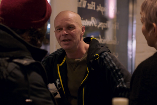 A bald white man wearing a hoodie talks to a small film crew