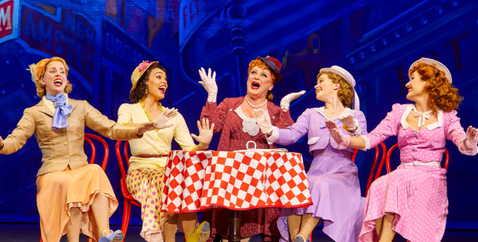 5 women in colourful tea dresses sit around a table with a red gingham table cloth on it