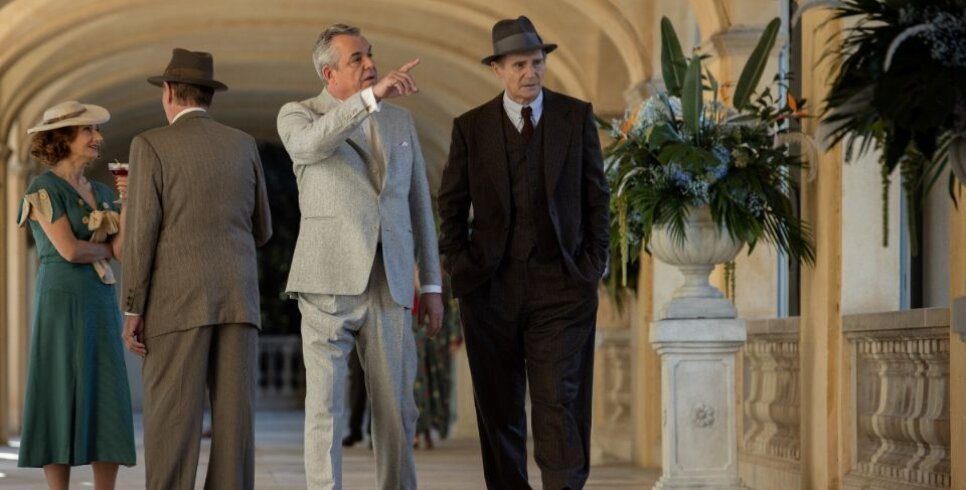 Liam Neeson (right) is playing a detective and is wearing a black three piece suit with a hat while a man in a grey suit is on his left pointing to something on the right out of sight. There are some other people behind them to their left wearing 30s style outfits.