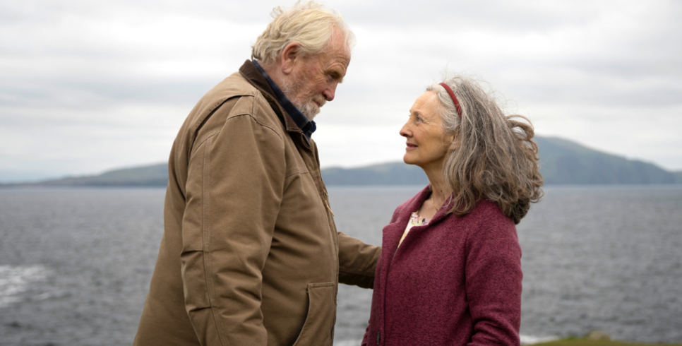On the left there is an old man in a beige coat looking an old woman (right) wearing a burgundy coat. They both have grey hair and are looking at each other lovingly. In the background there the sea and mountains.