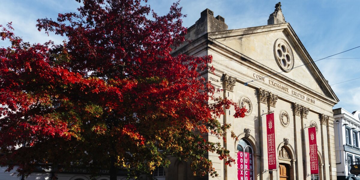 A view of the front of the Corn Exchange, an old building originally built in 1861 with a light stone facade. There is a tree to the left of it, the leaves red with autumn colour.