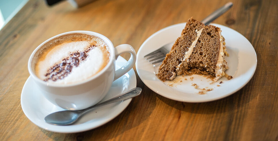 On a wooden surface is a coffee in a white mug with a saucer. On top of the coffee is the Corn Exchange logo, dusted in chocolate. To the right is a slice of coffee cake on a white plate with a fork.
