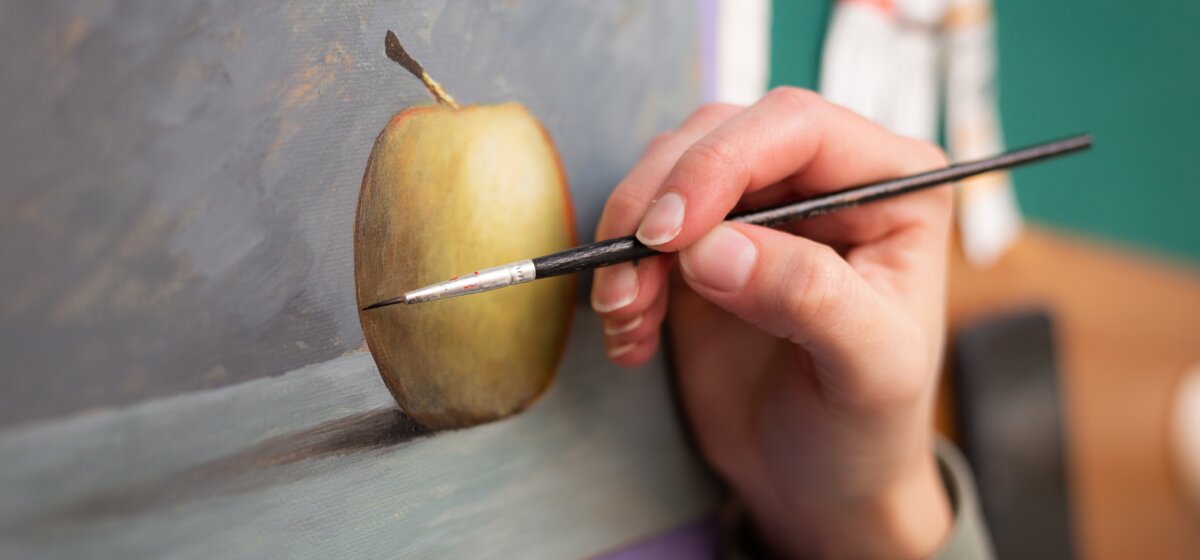 A hand holds a paintbrush and paints a picture of an apple.