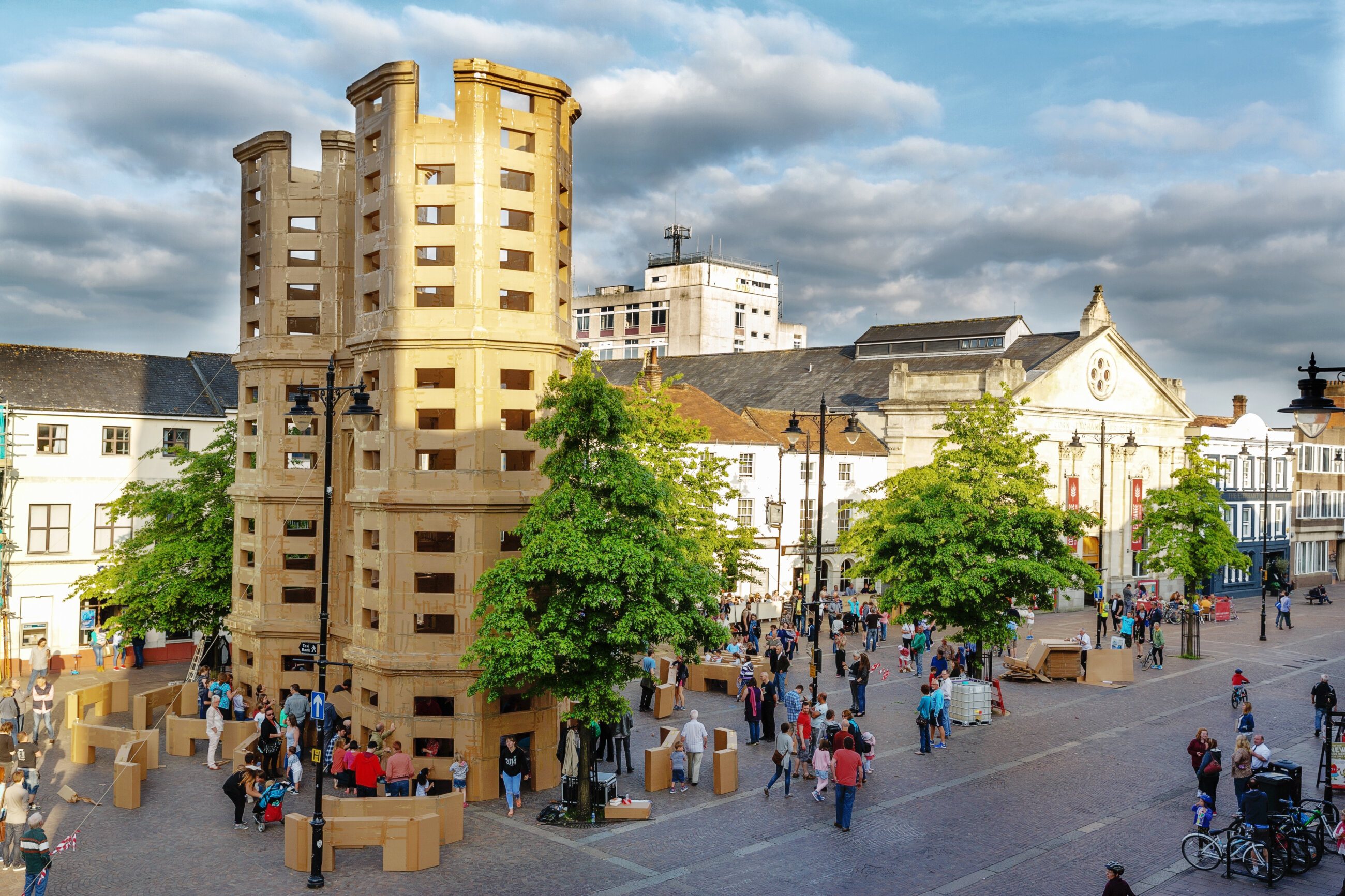 Image is of a large cardboard building in Newbury market place. The cardboard building is larger than all the other real buildings that surround it. Public audiences gather is small groups to admire the vast size of the 'building' structure constructed out of cardboard boxes.