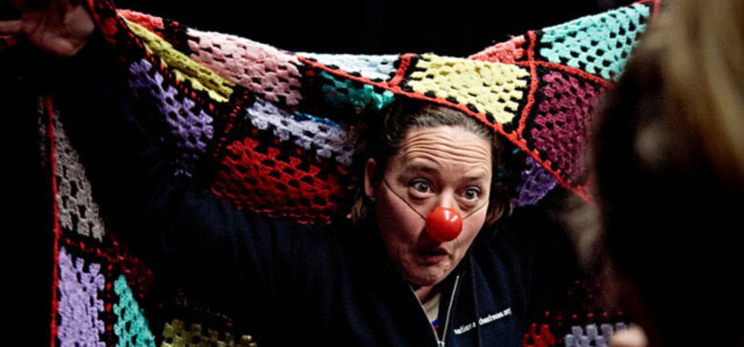 A person with a red nose makes a clown like facial expression and holds a crocheted blanket up behind them.