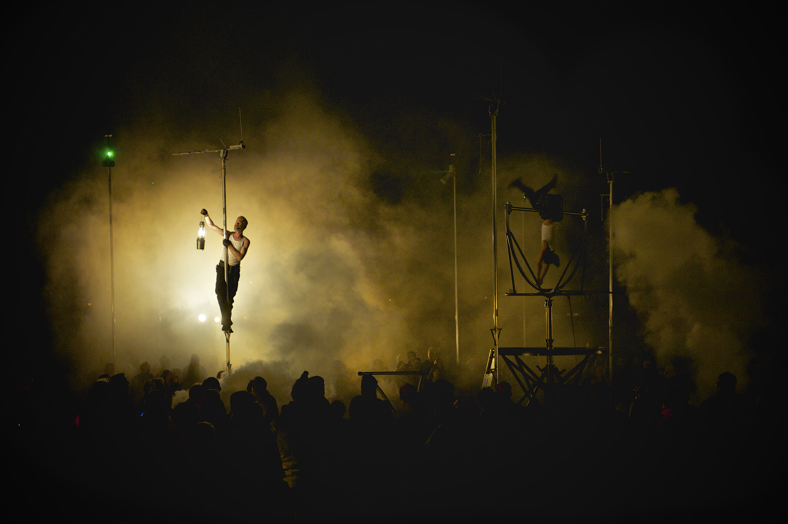 This image is of 2 artists performing at an outdoor show with an audience. One of the artists is standing on a metal pole, holding a lamp lantern to the left.  The other artist to the right of the image is caught mid somersault with both legs in the air while they perform on a different metal structure. The image is taken at night and the sky is lit up with an orange ambient smoke/mist.