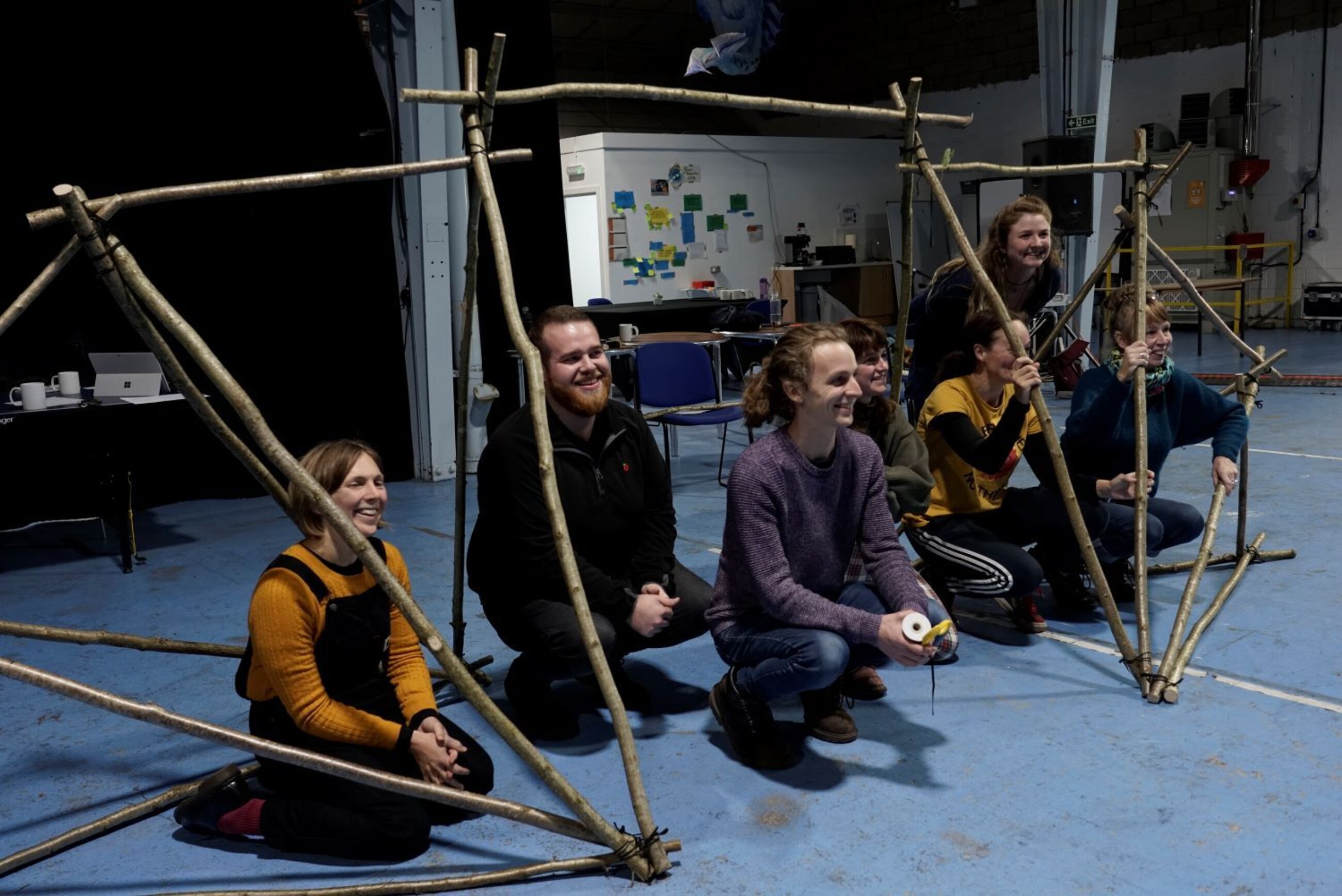 A group of people pose squatted in between structures that they've built out of branches and string.