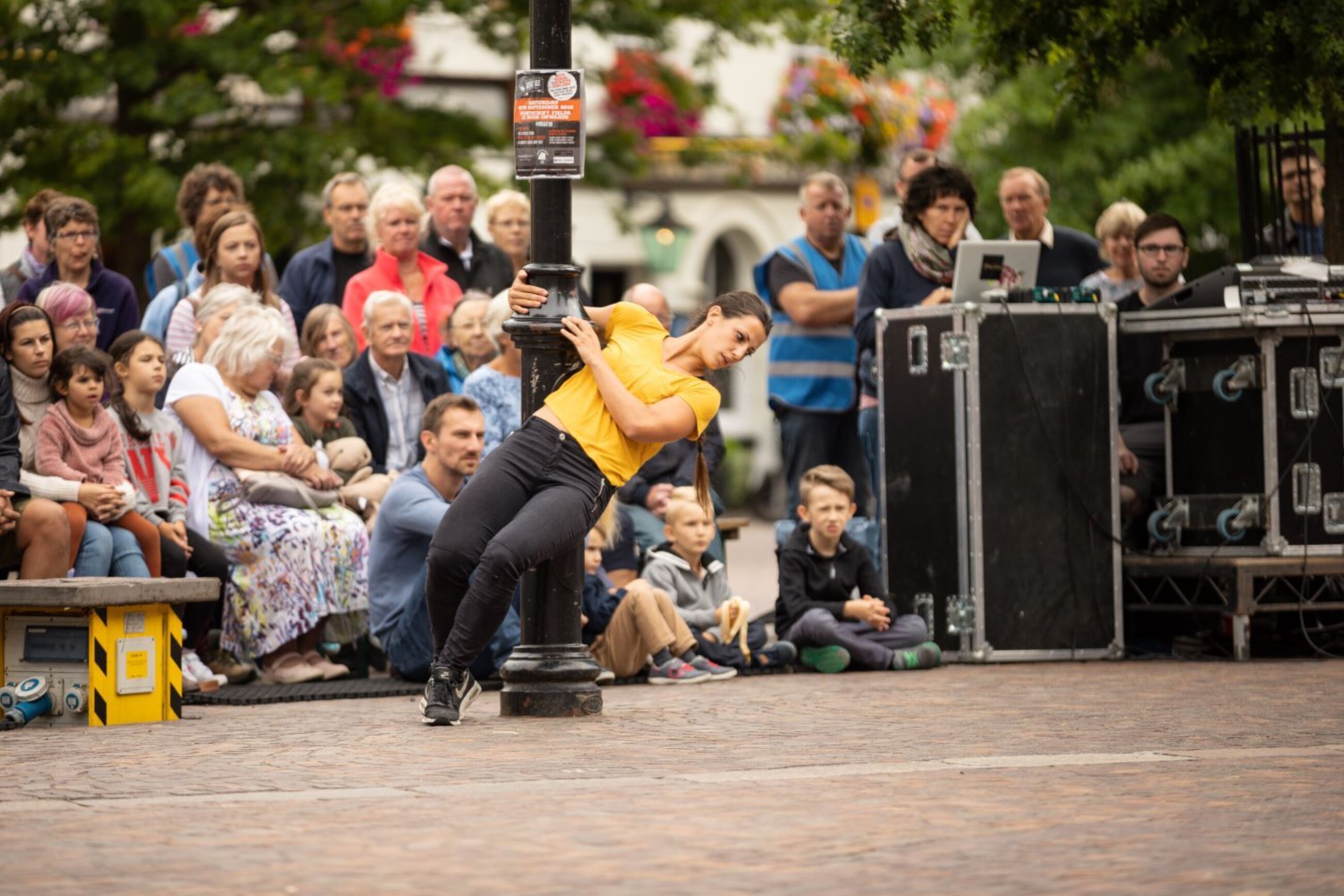 A woman in a yellow t shirt and black trousers leans from a lamppost in front of a large outdoor mixed age audience in Newbury Market Place