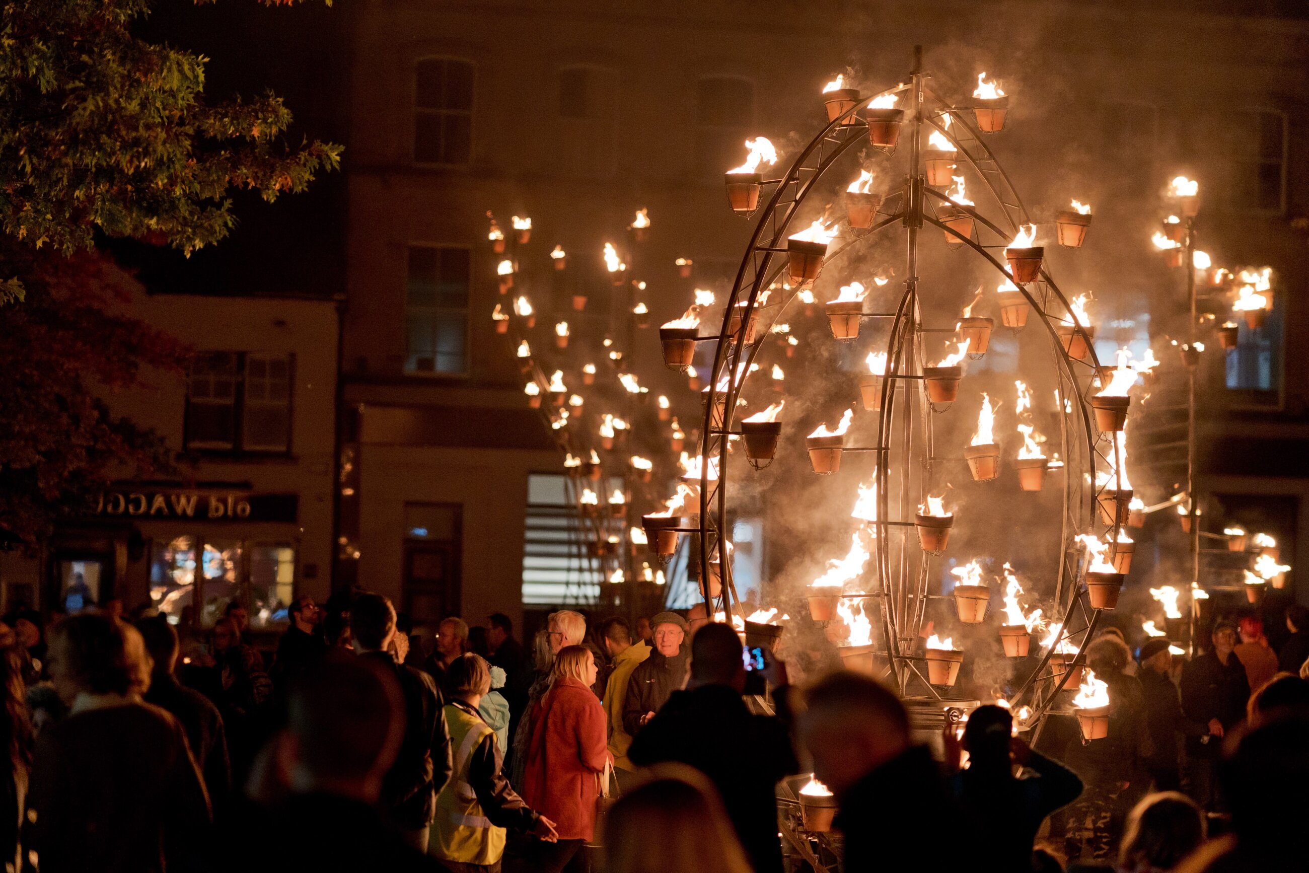 Image is of a public fire show hosted in Newbury market place. The image is at night time with an audience admiring a large metal framed wheel that has small ceramic plant pots placed around it. In each pot a small fire which lights up the circular framed structure.