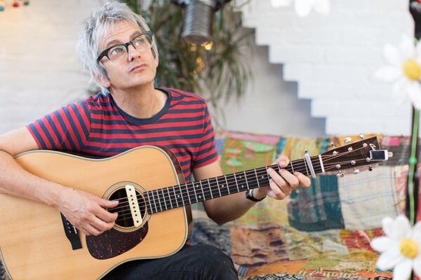 An image of Nick Cope sat on a patchwork sofa, looking thoughtful and holding his guitar