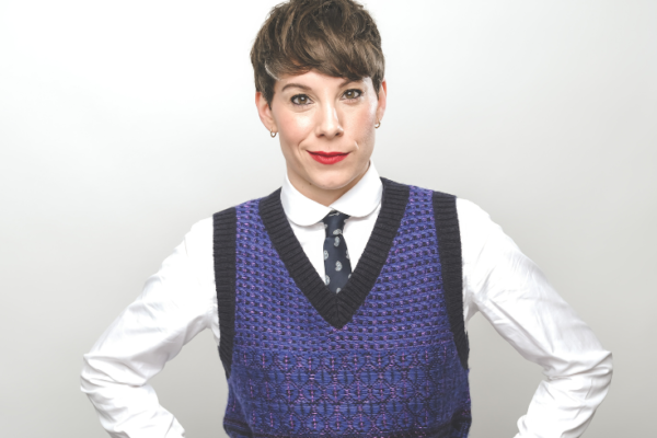 An image of comedian Suzi Ruffnell in a white shirt, blue knitted vest and tie coyly smiling to the camera with her hands on her hips.