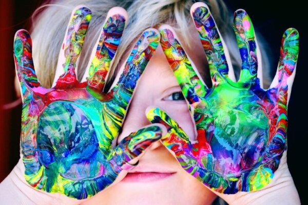 A photo of a young boy with his hands up covering his face aside from one eye and his lips. On his hands there is colourful marbled paint.