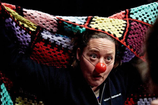 A person with a red nose makes a clown like facial expression and holds a crocheted blanket up behind them.