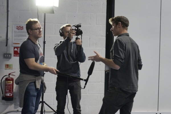 Image of 3 people. One talking with a mic, one holding mic and one filming with a hand held camera