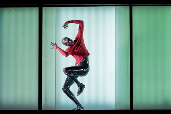 An alien like figure in a silver bodysuit wearing a red jumper strikes a dance like pose, behind a glass window lit by green light.
